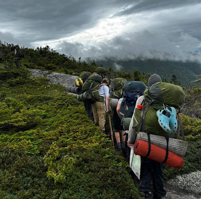 A group of hikers is trekking up a mountain trail, each carrying large backpacks. The path is surrounded by lush green vegetation, and the sky is overcast with clouds. The hikers are walking in a single file, suggesting a sense of camaraderie and shared adventure. The overall scene conveys a feeling of exploration and connection with nature.
